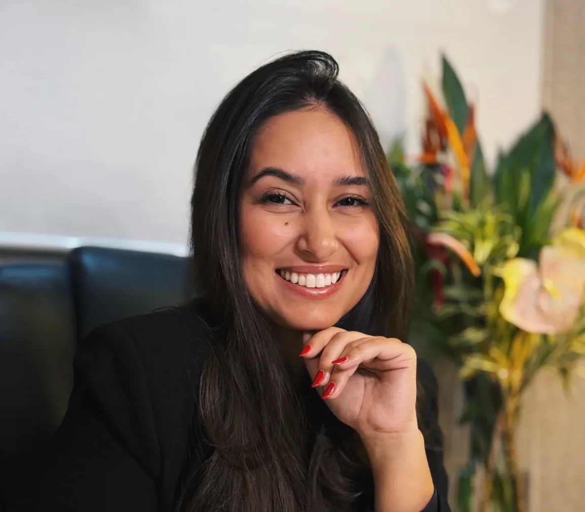 Mulher sorridente de blazer preto, sentada em um sofá de couro, com um arranjo de flores tropicais ao fundo